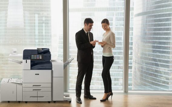 Deux personnes en tenue professionnelle, dans un bureau, discutent de documents, debout près d'une imprimante multifonction Xerox® AltaLink® série B8200.
