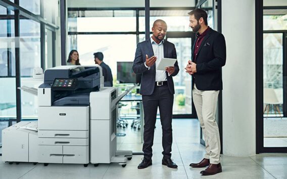 Deux hommes dans un bureau, en train de discuter devant une tablette, à côté d'une imprimante multifonction couleur Xerox® AltaLink® série C8200