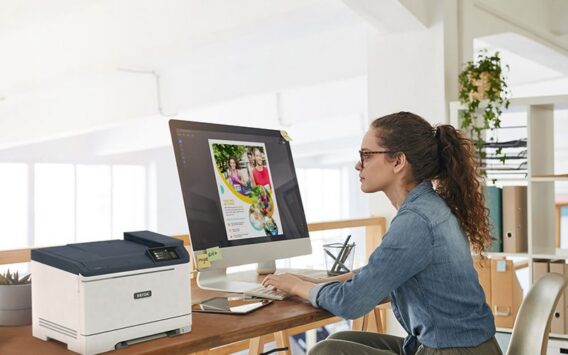Une femme est assise à un bureau, concentrée sur son travail, avec une imprimante couleur Xerox® C320 à ses côtés.